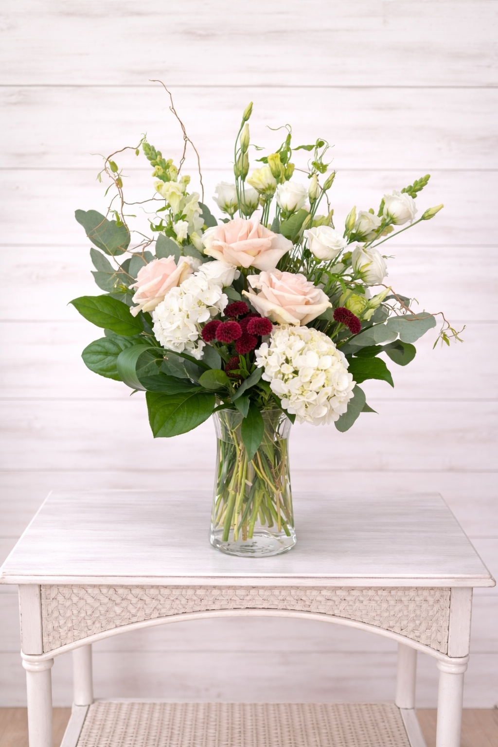Bouquet of white, pink, and burgundy flowers in a clear vase on a white table with a white wooden background