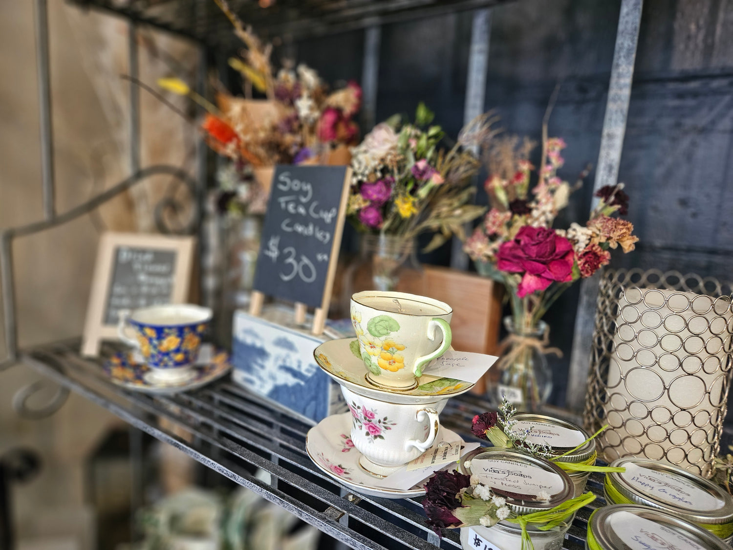 Teacups candles and dried flowers on a metal shelf with flowers and signs in the background