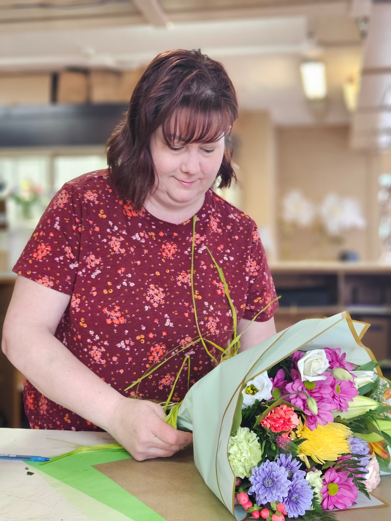 A woman in a red shirt putting together a bouquet of flowers in an indoor setting